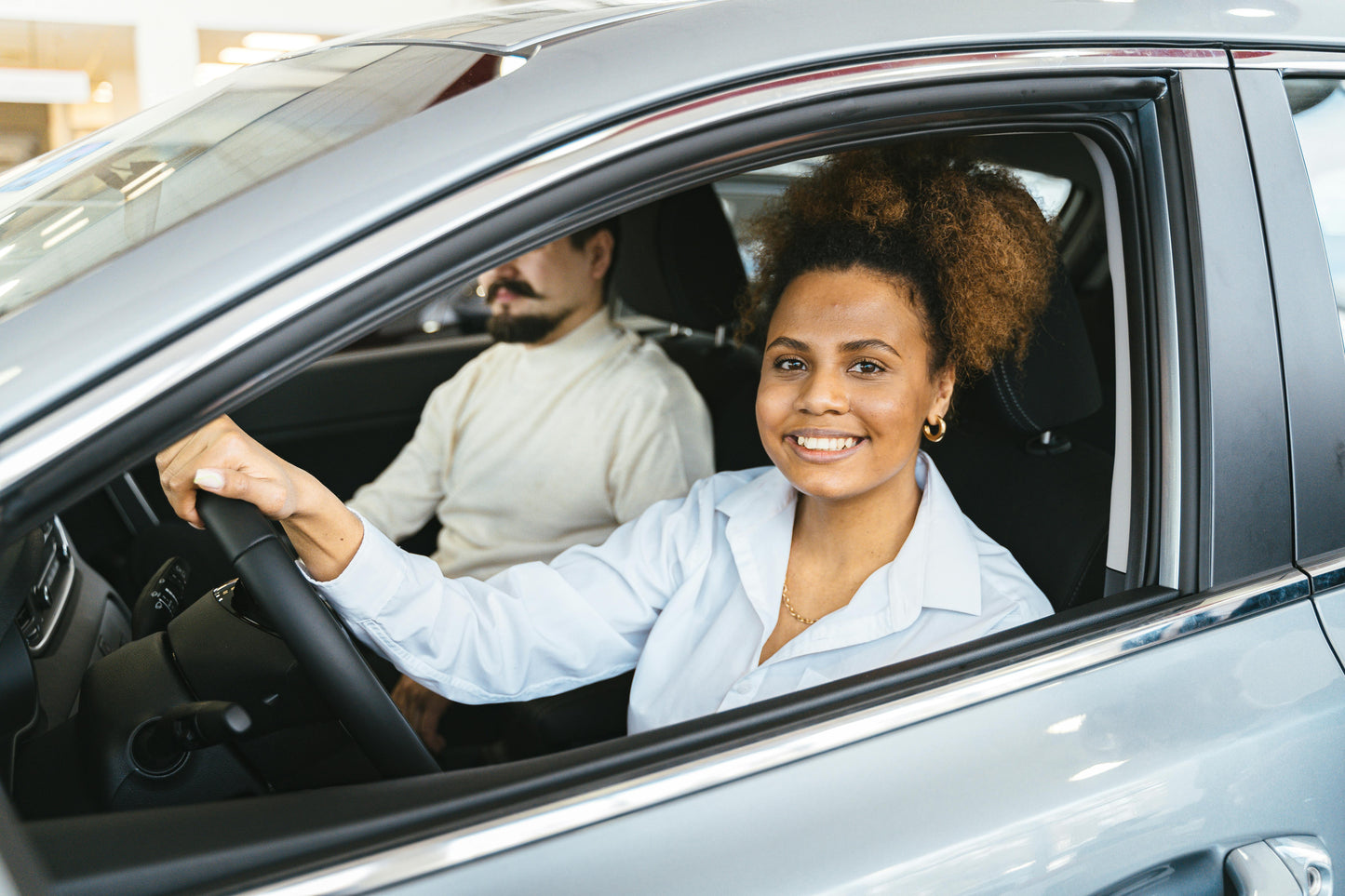 A young female student sits in the driver’s seat of a car, attentively listening to her driving instructor beside her. The instructor, a professional and patient guide, gestures as he provides instructions, ensuring she gains confidence behind the wheel.