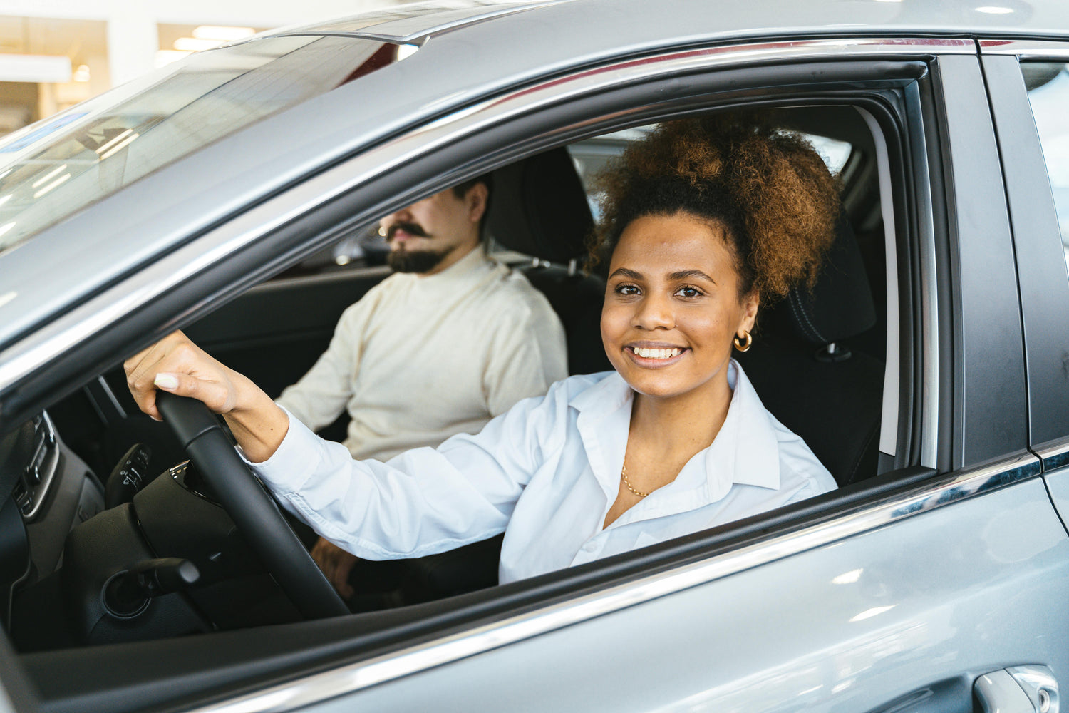 A young female student sits in the driver’s seat of a car, attentively listening to her driving instructor beside her. The instructor, a professional and patient guide, gestures as he provides instructions, ensuring she gains confidence behind the wheel.